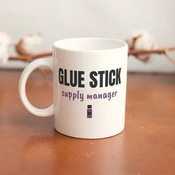 White 11 oz ceramic mug printed with "Glue Stick Supply Manager" on a wooden table