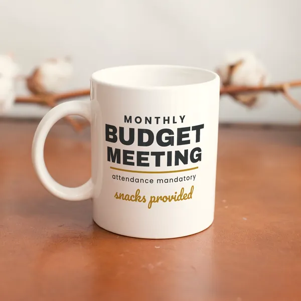 White 11 oz ceramic mug printed with "Monthly Budget Meeting - Snacks Provided" on a wooden table