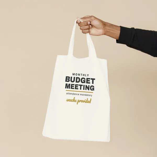 Natural canvas tote bag printed with "Monthly Budget Meeting - Snacks Provided", held by the handles