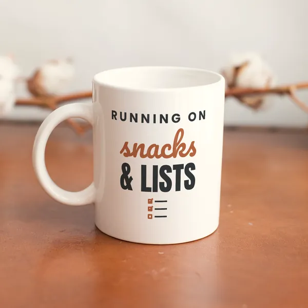 White 11 oz ceramic mug printed with "Running on Snacks and Lists" on a wooden table