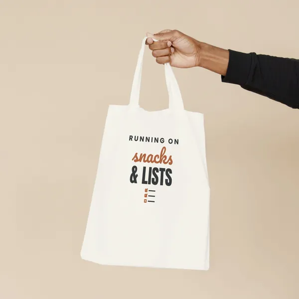 Natural canvas tote bag printed with "Running on Snacks and Lists", held by the handles